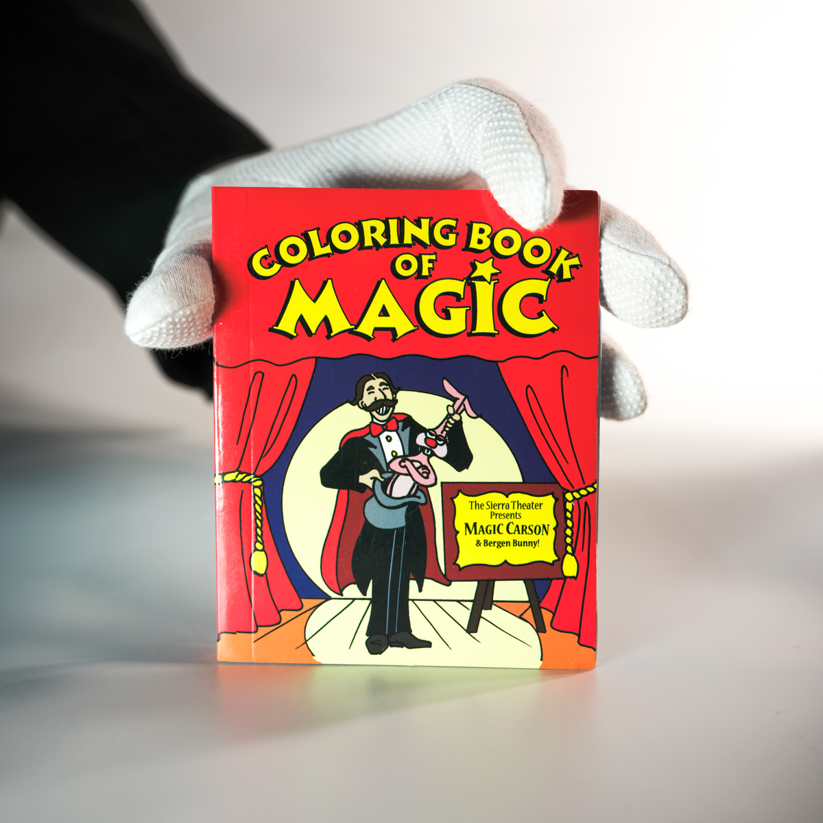 A gloved hand holds the Magic Coloring Book - Pocket Size 4 X 5 in, ideal for beginner magicians, showing an illustrated magician pulling a rabbit from a hat on stage.